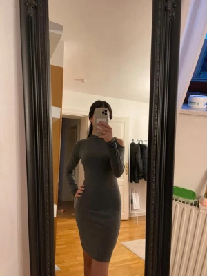 Mörkgrå tight klänning - Säljer en mörkgrå, tight klänning med lång ärm och trendig cut out-detalj på axeln. Klänningen har en hög hals och är i mjukt, stretchigt material som sitter snyggt på kroppen. Perfekt för dig som gillar minimalistisk och stilren look.