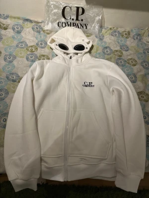 Vit hoodie från C.P. Company - Säljer en vit hoodie från C.P. Company med dragkedja och ikoniska glasögon i huvan. Helt ny. Med tags på. Pris går att diskuteras. Öppen för frågor och bud. Mått kan skickas