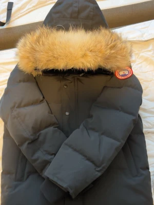 Svart dunjacka från Canada Goose - Säljer en svart dunjacka från Canada Goose med stor huva och fluffig pälsdetalj. Jackan har en klassisk patch på ärmen och är perfekt för kalla vinterdagar. Fylld med dun för extra värme och har en stilren, tidlös look.
