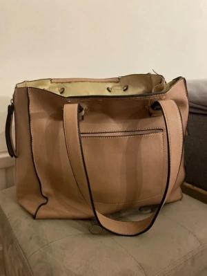 Ljusrosa handväska med svarta detaljer - Stilren ljusrosa/beigehandväska i konstläder med svarta kanter och dubbla handtag. Väskan har en praktisk ytterficka och ljus insida med tryckknappar. Perfekt för dig som vill ha plats för allt du behöver och gillar enkel, modern design.