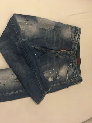 Blå slitna jeans med detaljer - Säljer ett par blå jeans från Dsquared2 med slitningar, hål och färgstänk för en riktigt edgy look. Jeansen har klassisk femficksmodell och knappgylf. Perfekta för dig som gillar streetstyle och vill sticka ut lite extra.