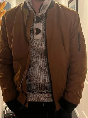 Brun bomberjacka med fickdetaljer - Snygg brun bomberjacka med klassisk passform och svarta muddar vid ärmslut och midja. Jackan har en ficka med dragkedja på ena ärmen och stängs med dragkedja framtill. Perfekt för dig som vill ha en stilren och trendig look. Inga defekter, skriv vid frågor eller intresse🥰🥰