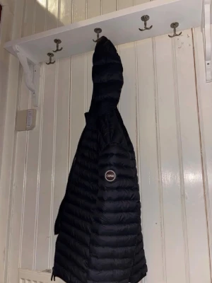 Svart pufferjacka från Colmar - Snygg svart pufferjacka från Colmar med huva och dragkedja framtill. Jackan har två praktiska fickor med dragkedja och ett Colmar-märke på ärmen. Lätt vadderad och perfekt för kalla vinterdagar. Klassisk design som funkar till det mesta. Sitter som S, passar dig som är mellan 165-178