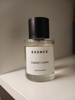 ESSNCE Sweet Cara Eau de Parfu💞🦋 - Minimalistisk parfymflaska från ESSNCE med doften Sweet Cara. Flaskan är cylindrisk i genomskinligt glas med en enkel vit etikett och svart plastlock. Sprayfunktion i metall. Passar dig som gillar stilren design och fräscha dofter.