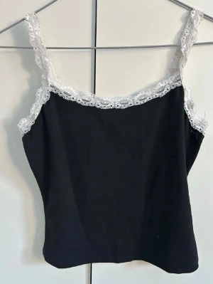 Svart linnetopp med spetskant - Snygg svart linnetopp med vita spetsdetaljer längs axelbanden och urringningen. Toppen är ärmlös och har en croppad passform som passar perfekt till jeans eller kjol. Materialet känns mjukt och stretchigt, vilket gör den bekväm att bära.
