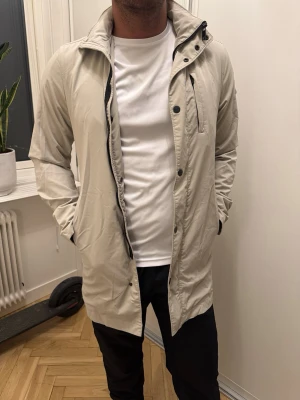 J.Lindeberg beige trenchcoat  - Stilren beige trenchcoat från J.Lindeberg med hög krage Jackan har en ogenerad bröstficka med dragkedja och två sidofickor. Liten svart patch på är pärmen. Lätt material, perfekt för lager-på-lager och som vårjacka. Nypris 4500 kr så gör ett kap. Jättefint skick 