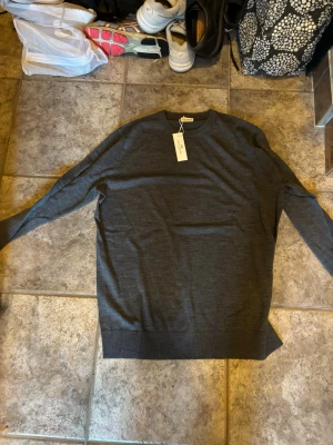 Mörkgrå stickad tröja John Henric  - Snygg mörkgrå stickad tröja från John Henric . Tröjan har en minimalistisk design och är tillverkad i ett tunt, mjukt merinoull som passar perfekt till lager på lager. Klassisk passform och ribbade muddar. Aldrig använd innan då den är helt för stor för mig. Storlek L