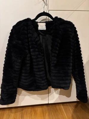 Svart pälsjacka från Jacqueline de Yong - Säljer en svart, fluffig pälsjacka från Jacqueline de Yong. Jackan har lång ärm, öppen framtill och ett stilrent, ribbat mönster i pälsen. Perfekt för dig som vill ha en trendig och mysig jacka till kyliga dagar.