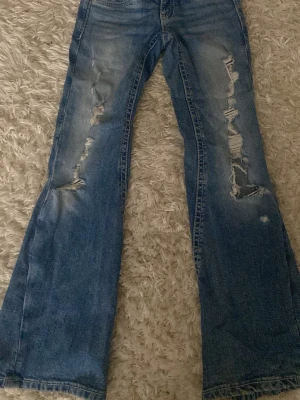 Blå slitna bootcut jeans Gina Tricot - Säljer ett par blå bootcut jeans från Gina Tricot med slitningar och hål på båda benen. Jeansen har en klassisk femficksmodell och är tillverkade i denim med snygg tvätt och slitna detaljer för en trendig look.