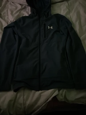 Mörk blå vindjacka från Under Armour - Snygg mörk blå vindjacka från Under Armour med huva och dragkedja framtill. Jackan har en diskret bröstficka med dragkedja och det klassiska Under Armour-loggan i vitt på bröstet. Tillverkad i lätt och tåligt material som står emot vind.