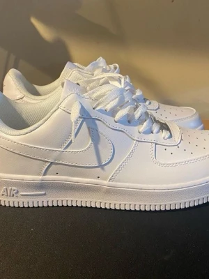 Nike Air Force 1 vita sneakers - Klassiska helvita Nike Air Force 1 sneakers i lågt utförande. Skorna har en stilren design med perforerad tå, kraftig sula och ikoniska swoosh-loggan på sidan. Tillverkade i slätt läder med matchande vita skosnören och detaljer.