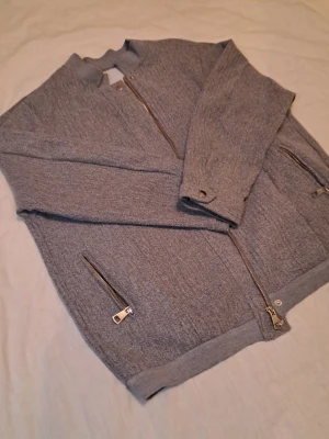 Grå bomberjacka med dragkedjor - Snygg grå bomberjacka i mjukt tyg med silverfärgade dragkedjor framtill och på fickorna. Perfekt för en stilren och avslappnad look. (Meddela  innan du köper!)