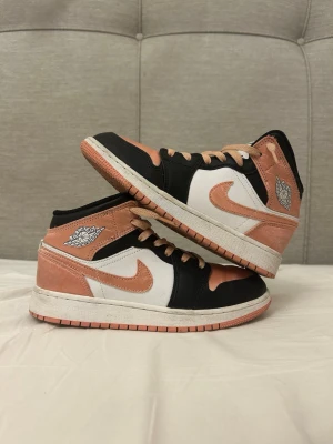Air Jordan 1 Mid Peach/Black/White - Snygga Air Jordan 1 Mid sneakers i färgerna persika, svart och vitt. Skorna har klassisk Jordan-logga på sidan och tungan, samt persikofärgade snören och swoosh. Ovandelen är i textil och läder för en cool och sportig look.