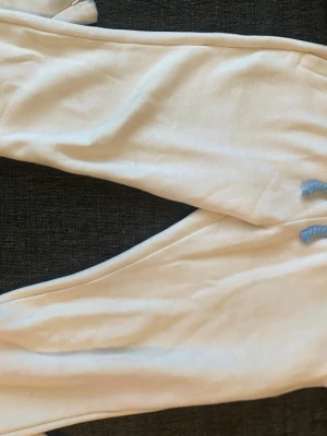 Syna World mjukisbyxor - Snygga vita mjukisbyxor med elastisk midja och blått graffiti-tryck på baksidan. Byxorna har dragsko i midjan och är tillverkade i mjukt material, perfekta för en avslappnad stil.