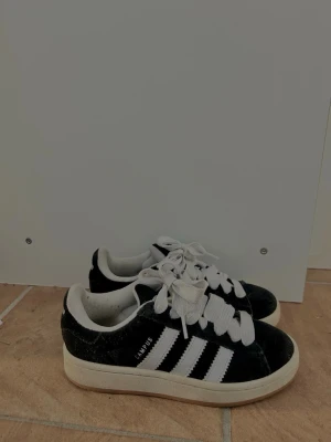 Adidas Campus  - Klassiska Adidas Campus sneakers i svart mocka med vita detaljer och de ikoniska tre ränderna på sidan. Använda 10 gånger som mest.