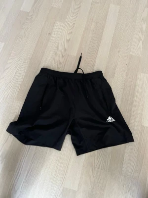 Svarta träningsshorts Adidas - Säljer ett par svarta träningsshorts från Adidas med snörning i midjan och logotyp på ena benet. Shortsen har en loose passform och är tillverkade i lätt syntetmaterial, perfekt för sport och aktivitet. Storleksmässigt hade jag sagt M