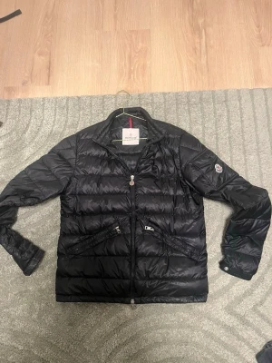 Svart dunjacka från Moncler - Snygg svart dunjacka från Moncler med klassisk quiltad design, dragkedja framtill och två fickor med dragkedja. Jackan har en liten Moncler-logga på ärmen och diskreta detaljer som ger en clean look. Perfekt för kalla vinterdagar.