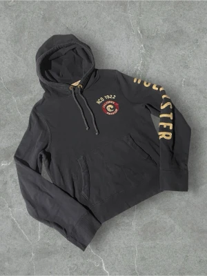 Mörkblå hoodie från Hollister | 38/M - Svart hoodie från Hollister med broderad logga och texten 'HCO 1922 California Beach & Surf' på bröstet. Stor gul text 'HOLLISTER' längs ena ärmen. Klassisk känguruficka och justerbar huva med snören. Mjuk bomullskänsla, perfekt för chill dagar.