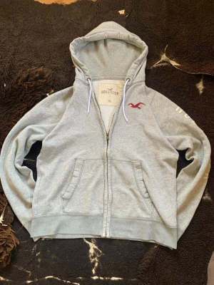 Grå zip hoodie från Hollister - Snygg grå hoodie från Hollister med dragkedja, två fickor fram och vit dragsko i huvan. Insidan är mjukt fodrad med teddy och loggan i rött broderi på bröstet. Skriv vid funderingar! Svarar snabbt.