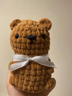 Brun virkad nalle med rosett - Söt handvirkad nallebjörn i mjukt, brunt garn med svart nos och ögon. Nallen har en vit sidenrosett runt halsen och små runda öron. Perfekt som dekoration eller present till någon som gillar gulliga saker.