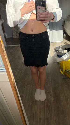 Svart jeanskjol med rå kant - Säljer en svart kort jeanskjol med rå, fransad kant nertill och klassiska fickor fram och bak. Kjolen har normal passform och stängs med knapp och dragkedja. Perfekt till en avslappnad och trendig look.