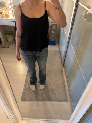 Svart spetslinnetopp med tunna band - Snygg svart linnetopp med tunna axelband och spetsdetaljer. Perfekt att styla med jeans eller kjol för en trendig look. Luftig och lätt, passar bra till varma dagar.