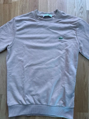 Beige sweatshirt från Lacoste - Säljer en stilren beige sweatshirt från Lacoste med den klassiska gröna krokodilloggan broderad på bröstet. Tröjan har rund halsringning, ribbade muddar och är tillverkad i mjukt bomullsmaterial. Perfekt för en avslappnad och trendig look.