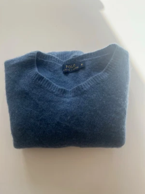 Blå stickad tröja Polo Ralph Lauren - Mysig blå stickad tröja från Polo Ralph Lauren med v-ringning och ribbade muddar vid ärmslut och nederkant. Tillverkad i ett mjukt material som ger en avslappnad och stilren look. Perfekt för lager på lager under kyliga dagar. Har krympt lite i tvätten så den sitter mer som XS-S men annars är den som ny. Den är lite längre i armarna 