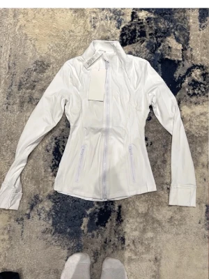 Vit träningsjacka  - Snygg vit träningsjacka  med figurnära passform, hög krage och hel dragkedja framtill. Jackan har två fickor med dragkedja och långa ärmar med tumhål. Perfekt för gymmet eller löprundan.