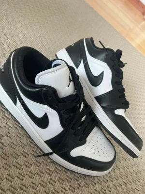 Air Jordan 1 Low svart/vit sneaker - Säljer ett par Air Jordan 1 Low sneakers i svart och vitt läder med klassisk Swoosh på sidan och Air Jordan-logga broderad på hälen. Skorna har svart snörning, perforerad tåbox och vit mellansula. Perfekta för dig som gillar streetwear och ikonisk design.