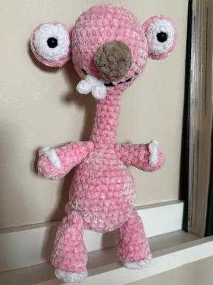 Virkad Sid från Ice Age mjukis - Unik handgjord virkad mjukisfigur som föreställer Sid från Ice Age. Figuren är rosa med vita och grå detaljer, stora ögon och lång hals. Tillverkad i mjukt garn, perfekt som dekoration eller present till någon som gillar filmkaraktärer.