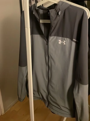 Grå träningsjacka från Under Armour - Snygg grå träningsjacka från Under Armour med dragkedja och huva. Jackan har två nyanser av grått, logga på bröstet och är tillverkad i ett lätt funktionsmaterial. Perfekt för löpning eller gym.
