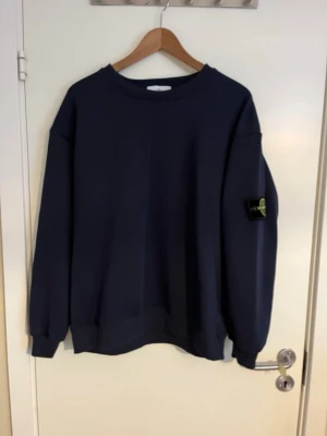 Mörkblå sweatshirt från Stone Island - Snygg mörkblå sweatshirt från Stone Island med klassisk rund hals och den ikoniska loggan på vänster ärm. Tröjan har en relaxed fit och ribbade muddar vid ärmslut och nederkant. Perfekt för dig som gillar streetwear och vill ha en stilren look.