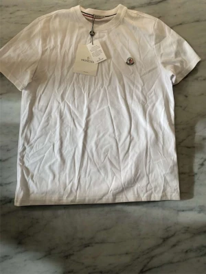 Vit Moncler t-shirt med logga - Snygg vit t-shirt från Moncler med klassisk rund hals och liten Moncler-logga broderad på bröstet. Tillverkad i mjuk bomull och har korta ärmar. Perfekt basplagg med exklusiv känsla och stilren design.