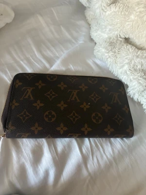 Louis Vuitton monogram plånbok - Säljer en klassisk brun plånbok från Louis Vuitton med det ikoniska monogrammönstret i guld. Plånboken har dragkedja runtom och är tillverkad i tåligt canvasmaterial med läderdetaljer. Perfekt för dig som gillar lyxiga accessoarer och vill ha plats för både kort och kontanter.