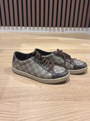 Gucci skor  - ÄKTA! Snygga låga sneakers från Gucci med klassiskt GG-monogram i beige och brunt. Skorna har bruna läderdetaljer, matchande bruna skosnören och vit sula. Perfekta för dig som gillar exklusiv och tidlös stil.