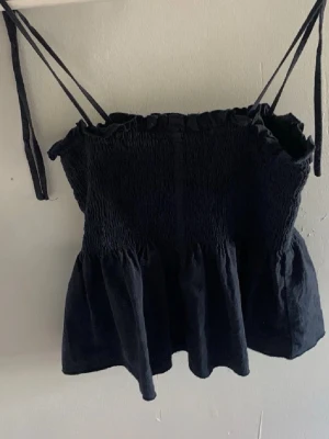 Blå smockad linnetopp med knytband - Trendig blå linnetopp med smockad överdel och volang nedtill. Tunna axelband som knyts upptill för en justerbar passform. Perfekt till jeans eller kjol för en cool och enkel look.