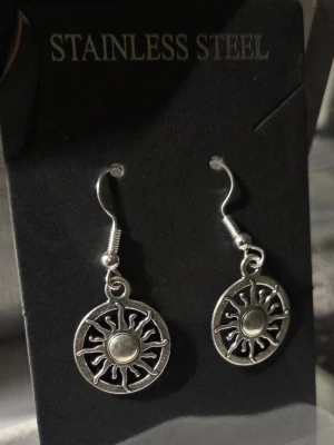 Solformade örhängen i rostfritt stål - Säljer ett par snygga örhängen i rostfritt stål med solmotiv. De är silverfärgade och har en rund form med detaljerade solstrålar. Perfekta för dig som vill ha en unik accessoar med cool vibe.