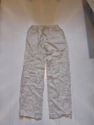 Beiga linnebyxor med dragsko - Säljer ett par beiga linnebyxor med avslappnad passform och dragsko i midjan. Byxorna har raka ben och är tillverkade i ett luftigt linnematerial, perfekta för varma dagar. Fickor i sidorna för extra funktionalitet.