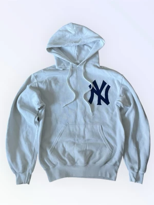 Vit hoodie med New York Yankees-logga - Snygg vit hoodie med stor mörkblå New York Yankees-logga på bröstet. Klassisk känguruficka framtill, justerbar huva med snören. Märke fruit of the loom. Några skavanker: lite nopprig, liten fläck på ryggen och små sprickor på loggan (se bild)