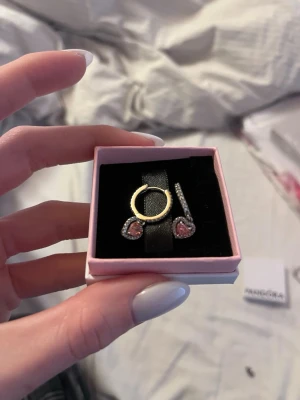 Pandora örhängen med rosa hjärta - Säljer ett par snygga örhängen från Pandora i silver med gnistrande stenar längs ringen och ett hängande hjärta i rosa. Hjärtat är dekorerat med små stenar som ger extra bling. Perfekt accessoar för dig som gillar romantiska detaljer och elegant stil. Aldrig använda. Nya kostar 1200kr