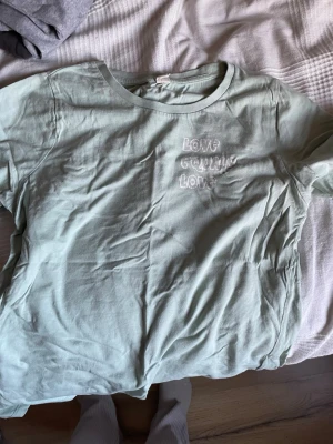 Ljusgrön t-shirt med broderi - Snygg ljusgrön t-shirt med broderad text 'LOVE GROWING LOVE' på bröstet. Klassisk rund halsringning och korta ärmar. Perfekt till jeans eller shorts för en chill look.