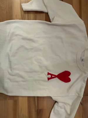 Vit stickad tröja från Ami - Vit stickad tröja från Ami Alexandre Mattiussi med röd hjärtlogga och bokstaven A på bröstet. Tröjan har rund halsringning och ribbade muddar vid ärmslut och nederkant. Perfekt för dig som gillar stilrena plagg med en twist.