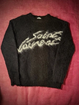 Svart fluffig tröja Saint Laurent - Stickad svart tröja från Saint Laurent med vit text i borstigt garn över bröstet. Tröjan har rund hals, långa ärmar och ribbade muddar. Materialet är mohair vilket ger en extra fluffig känsla. Skick som nytt!