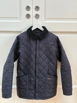 Mörkblå quiltad dunjacka från Barbour - Stilren svart dunjacka från Barbour med klassiskt rutquiltat mönster och svart manchesterkrage. Jackan har tryckknappar framtill, två stora framfickor och diskret Barbour-logga på fickan. Perfekt för våren eller hösten och ger en tidlös look. Jackan är i storlek Kids XL.