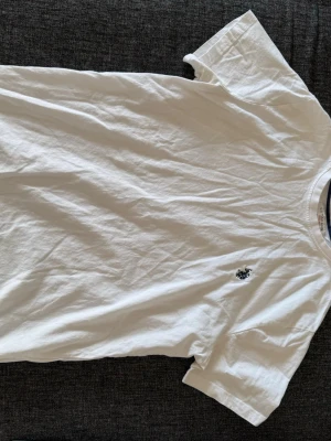 Vit t-shirt från U.S. Polo Assn - Klassisk vit t-shirt från U.S. Polo Assn med liten broderad logga på bröstet. T-shirten har rund halsringning med diskreta rödblå detaljer på insidan och är tillverkad i mjuk bomull. Perfekt basic-plagg för en clean och enkel stil.