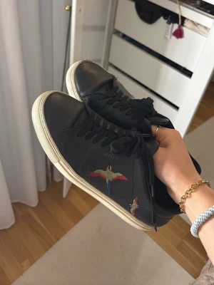 Svarta sneakers från Axel Arigato - Svarta sneakers från Axel Arigato med broderade färgglada fåglar på sidan. Skorna har vita sulor, rund tå och klassisk snörning. Tillverkade i skinn för en clean och stilren look som sticker ut med detaljerna. Säljer då de tyvärr är för stora