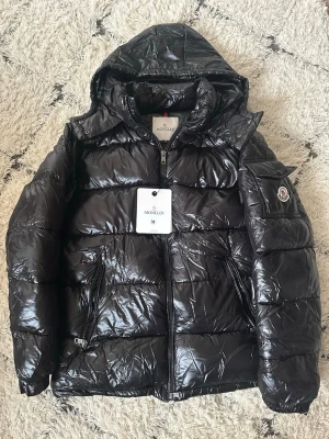 Moncler Maya  - Snygg svart dunjacka från Moncler med glansig finish, huva och dragkedja framtill. Jackan har två framfickor med dragkedja och Moncler-logga på vänster ärm. Perfekt för kalla vinterdagar och riktigt stilren look. Det är bara att höra av er om fler bilder önskas. Pris går alltid att diskuteras 🙏