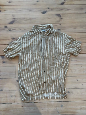 Beige randig skjorta Jack & Jones - Säljer en beige skjorta från Jack & Jones i storlek XL. Skjortan har vita vertikala ränder, korta ärmar och knappar framtill. Tillverkad i ett lätt och skönt material som passar perfekt till varmare dagar. Enkel och stilren design.