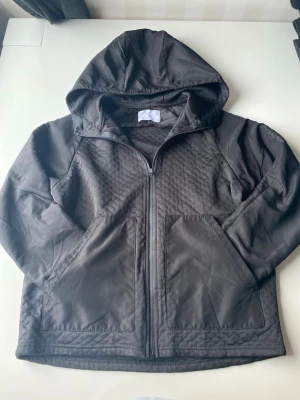 Svart hoodie med dragkedja från ZARA - Säljer en svart hoodie från ZARA med dragkedja framtill och två stora fickor. Hoodien har en klassisk huva och är tillverkad i ett mjukt, quiltat material som ger extra komfort. Perfekt för en avslappnad och stilren look.